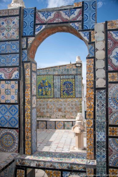 Large multicolored ceramic-tiled arch framing an open rooftop terrace and another patterned tile wall beyond.
