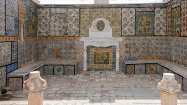 Small courtyard covered in patterned ceramic tiles, with a central white fountain niche, tiled benches, and stone pedestals.