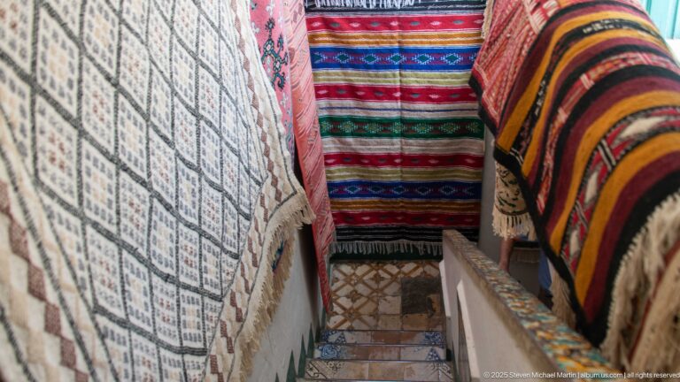 Narrow stairway lined on both sides with hanging woven rugs and blankets in red, black, cream, and multicolored patterns.