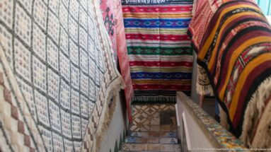 Narrow stairway lined on both sides with hanging woven rugs and blankets in red, black, cream, and multicolored patterns.