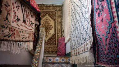 Vertical hanging rugs with geometric medallions and fringed edges fill a narrow passage, with another carpet visible at the far end.