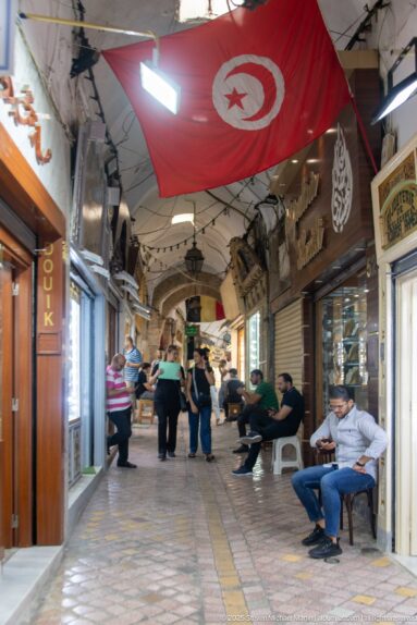 Covered market corridor with masonry arches, shopfronts, seated men, pedestrians, and a large Tunisian flag overhead.
