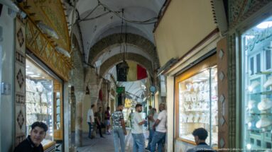 Narrow arcaded market corridor lined with brightly lit jewelry display windows, with shoppers and shopkeepers beneath rough masonry arches.
