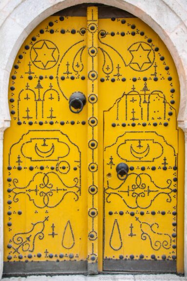 Close view of a yellow arched double door covered with symmetrical black motifs, studs, hinges, and circular bosses.