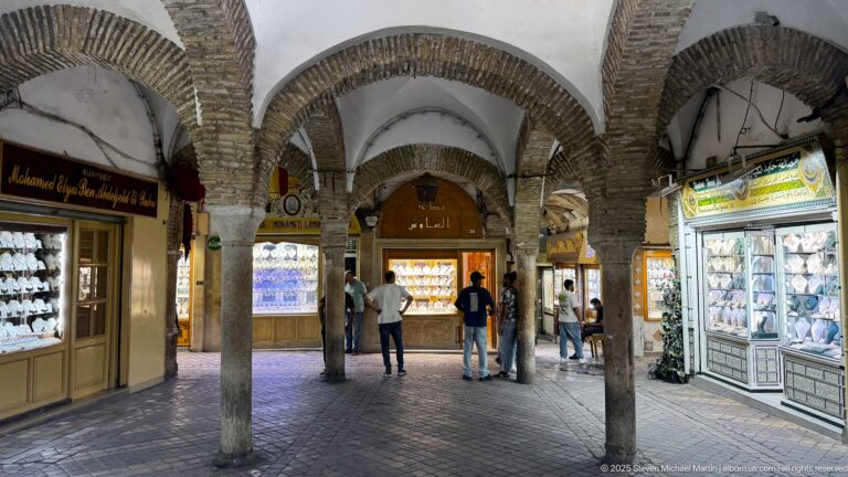 Arcaded jewelry market hall with stone columns, brick-edged arches, illuminated display cases, and several men in the central aisle.