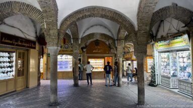 Arcaded jewelry market hall with stone columns, brick-edged arches, illuminated display cases, and several men in the central aisle.
