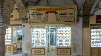 Jewelry shopfront beneath the arcade with French and Arabic signage and brightly lit cases filled with gold-colored necklaces and ornaments.