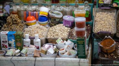 Market counter densely arranged with dried plant material, jars, bottles, soaps, boxed products, and small packaged goods.