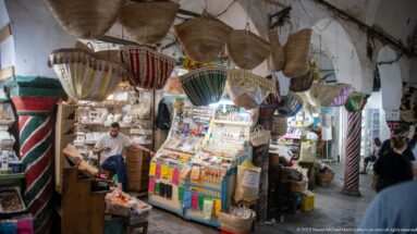 Compact market stall with hanging woven baskets, striped containers, shelves of small goods, and a seated vendor behind a brightly lit counter.