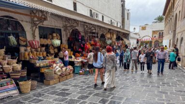 Busy stone-paved market lane with baskets, textiles, awnings, and numerous pedestrians between shopfronts and historic façades.