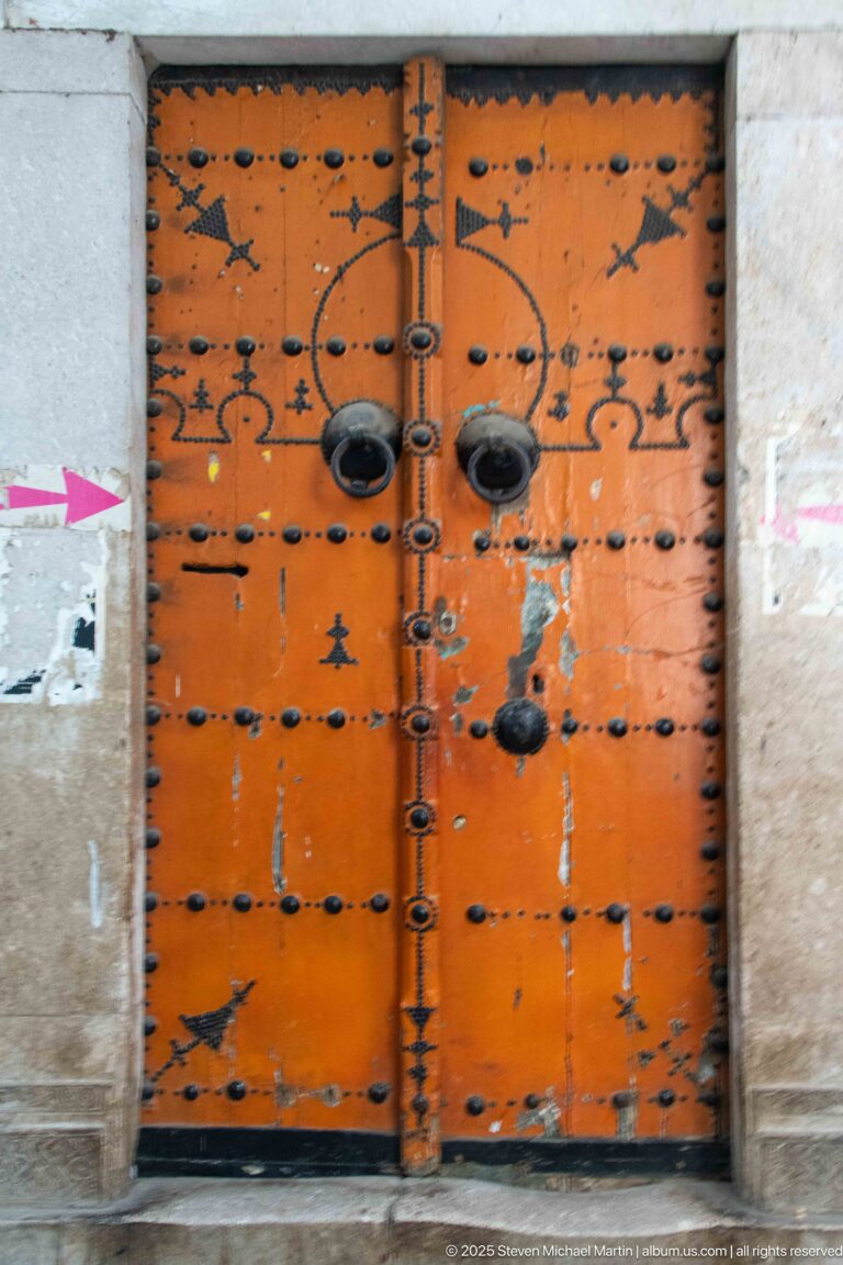 Weathered orange wooden double door with rows of black iron studs, paired projecting knockers, and worn paint.