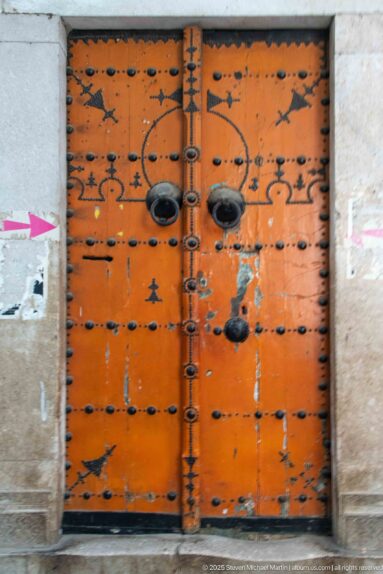 Weathered orange wooden double door with rows of black iron studs, paired projecting knockers, and worn paint.