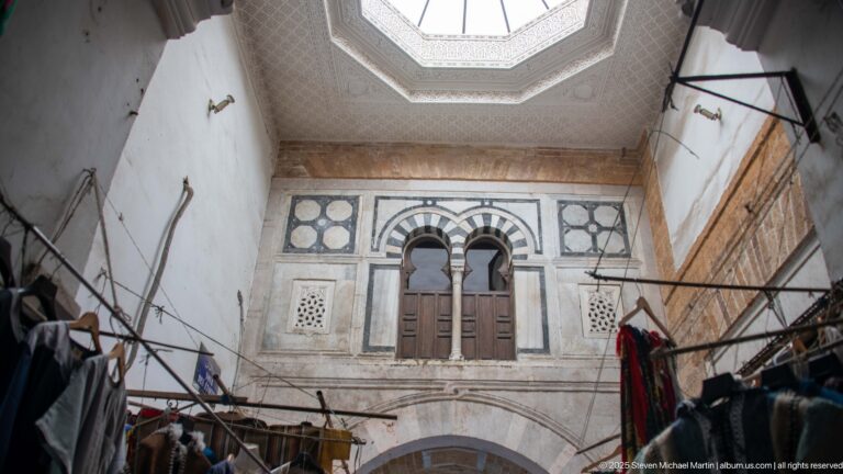 Skylit interior courtyard with paired arched upper windows, wooden shutters, carved panels, and black-and-white masonry bands above market fixtures.