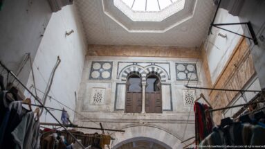 Skylit interior courtyard with paired arched upper windows, wooden shutters, carved panels, and black-and-white masonry bands above market fixtures.
