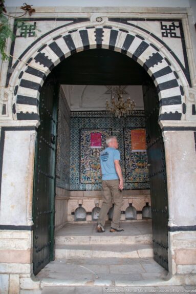 Broad black-and-white striped masonry arch with open doors framing a decorated interior and a visitor crossing the threshold.