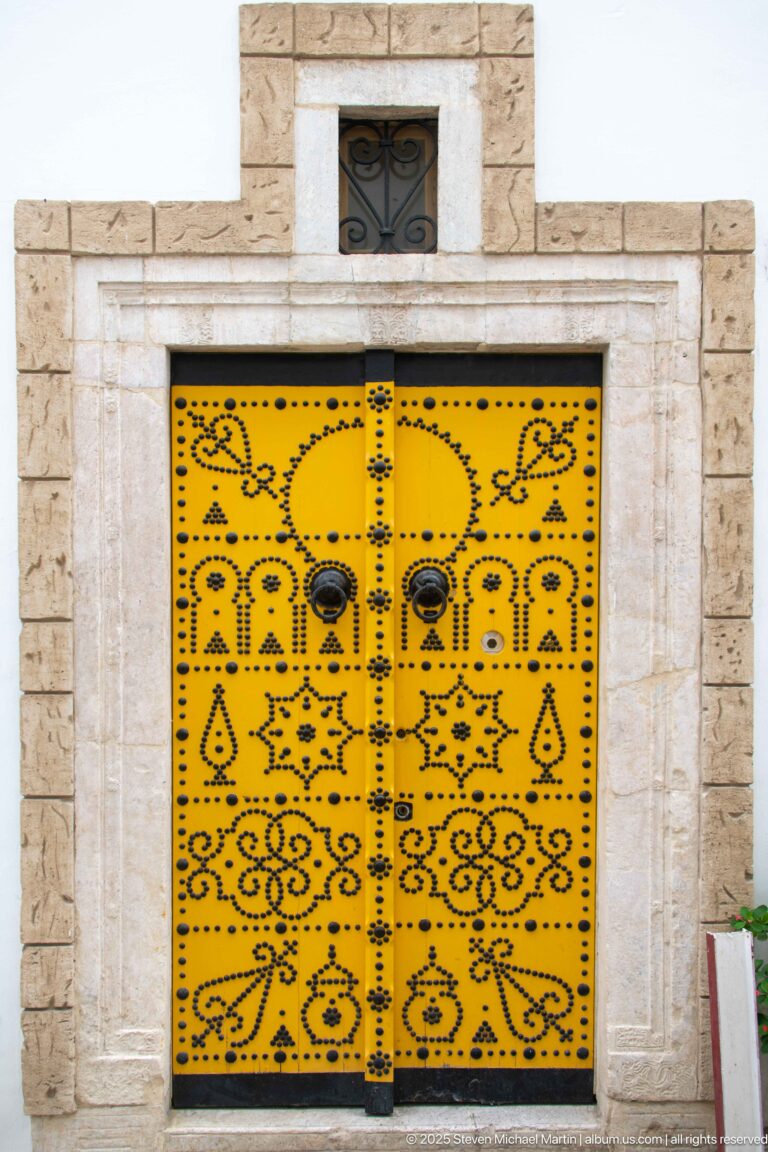 Yellow rectangular double door in a pale stone frame, decorated with black floral and geometric motifs and round metal bosses.