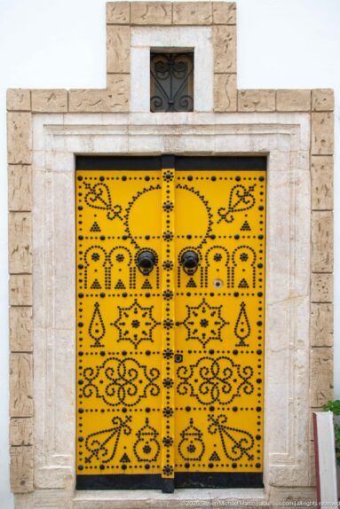 Yellow rectangular double door in a pale stone frame, decorated with black floral and geometric motifs and round metal bosses.