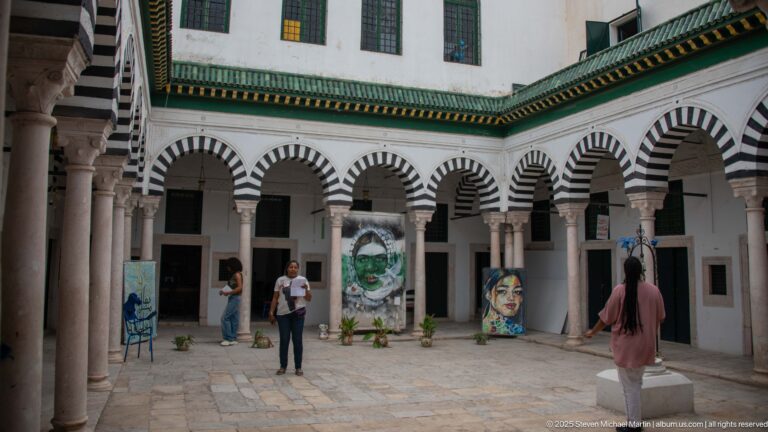 Courtyard surrounded by black-and-white striped arches and pale columns, with contemporary paintings and sculptural displays arranged in the open center.