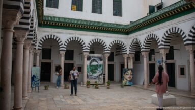 Courtyard surrounded by black-and-white striped arches and pale columns, with contemporary paintings and sculptural displays arranged in the open center.