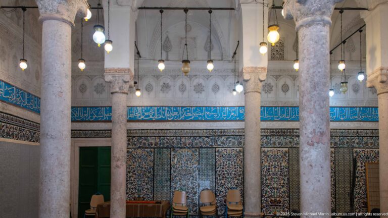 Pale stone columns and hanging lamps in a tiled hall with a turquoise Arabic calligraphic frieze and green doorway.