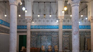 Pale stone columns and hanging lamps in a tiled hall with a turquoise Arabic calligraphic frieze and green doorway.