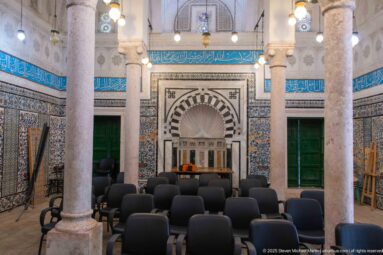 Rows of chairs facing an ornate striped-arch niche in a tiled hall with stone columns, green doors, and Arabic calligraphy.
