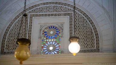 Small multicolored glass window surrounded by intricate white pierced plaster lattice beneath a larger arch.