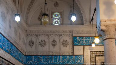 Turquoise Arabic calligraphic band below carved white plaster panels and a small colored-glass window, with hanging lamps nearby.