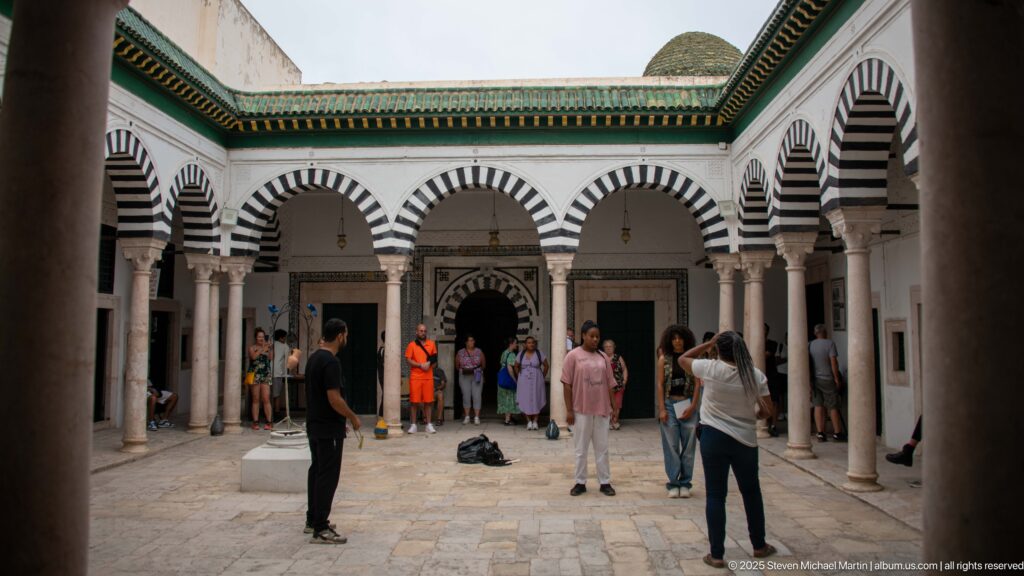 Open courtyard surrounded by striped horseshoe arches on stone columns, with green roof trim and groups of visitors on the paving.