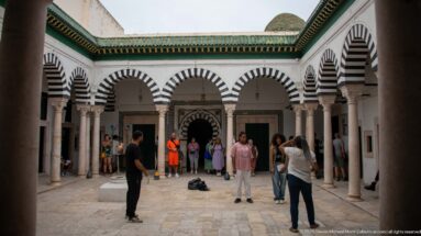 Open courtyard surrounded by striped horseshoe arches on stone columns, with green roof trim and groups of visitors on the paving.
