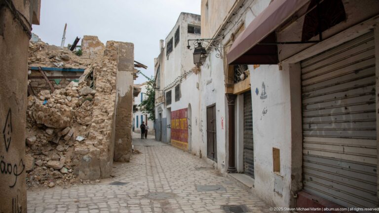 Narrow cobbled medina lane with closed shop shutters at right and a partially collapsed stone-and-rubble wall at left.