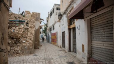 Narrow cobbled medina lane with closed shop shutters at right and a partially collapsed stone-and-rubble wall at left.