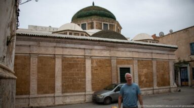 Low stone religious building with several white domes and a large green-tiled dome above an ochre façade divided by pale pilasters.