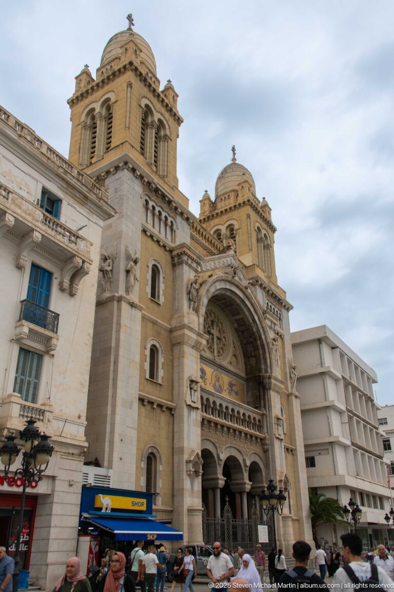 Oblique street view of St. Vincent de Paul Cathedral in Tunis, with twin towers, arched façade, neighboring buildings, and pedestrians.