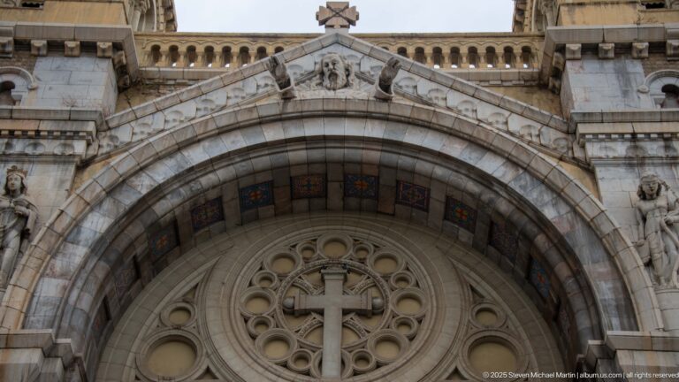Large cathedral entrance arch with colored soffit panels, circular cross-centered tracery, and sculpted gable above.