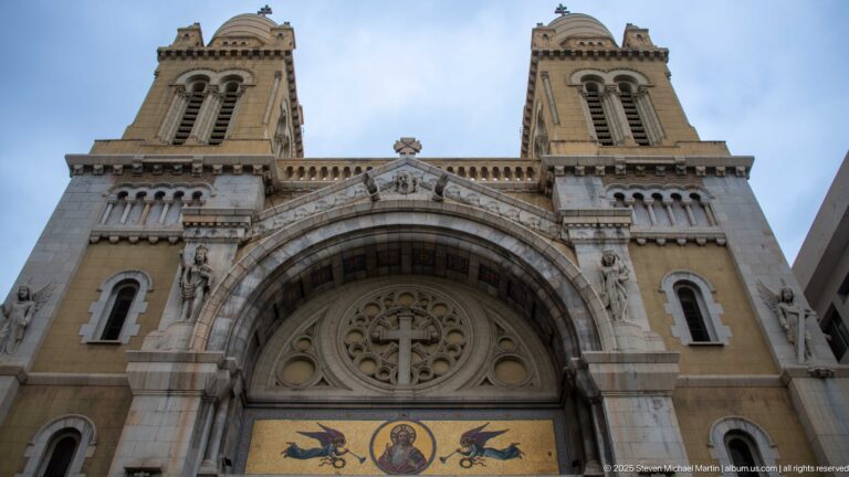 Low-angle cathedral façade with twin towers, circular tracery, and gold mosaic panel beneath the monumental entrance arch.