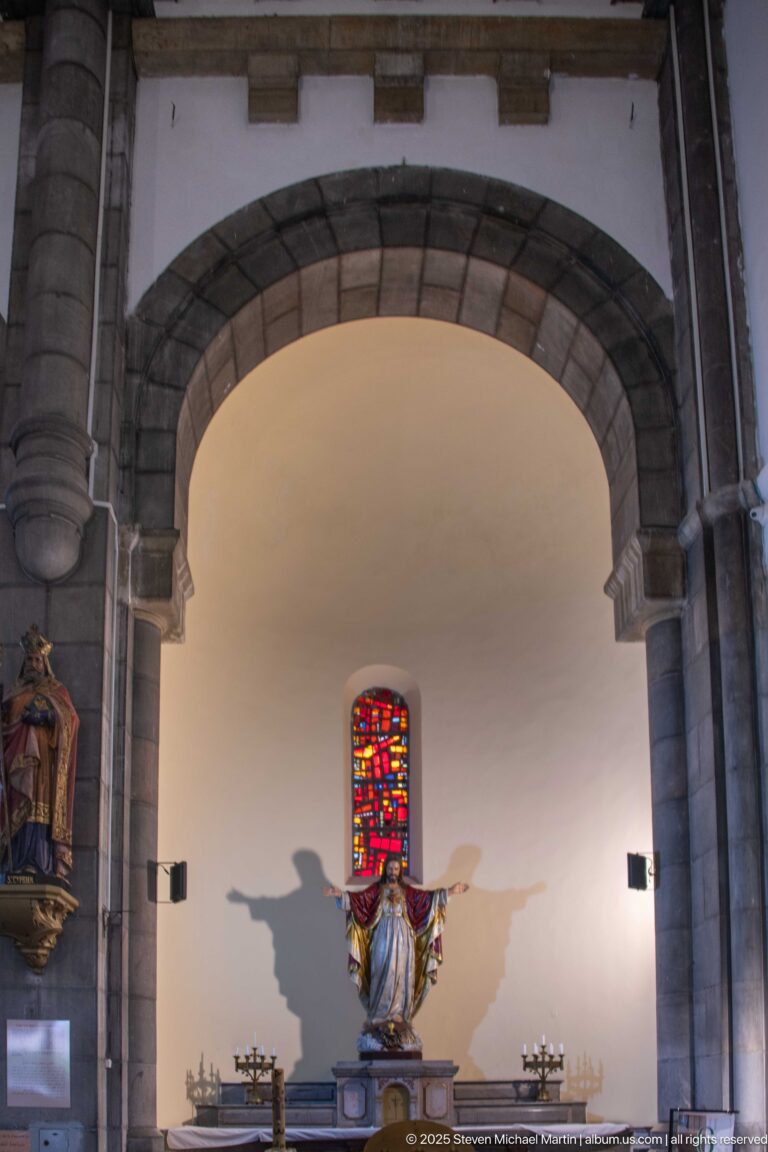Stone arch framing a side chapel with a polychrome statue of Christ, narrow stained-glass window, candles, and a second statue.