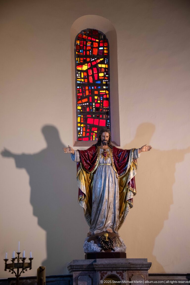Polychrome statue of Christ with outstretched arms beneath a narrow red stained-glass window, casting a large wall shadow.