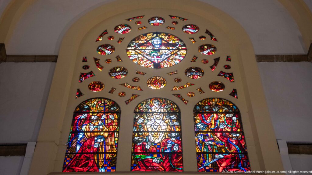 Large red-dominant stained-glass window with three figurative lower panels, central roundel, and many small surrounding openings.