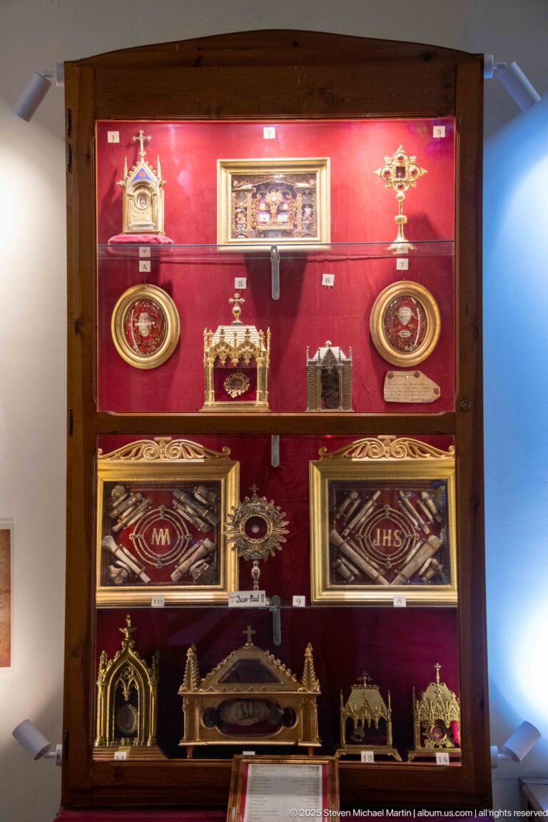 Glass display case with Catholic devotional objects, crosses, framed emblems, small shrines, medallions, and reliquary-like containers.