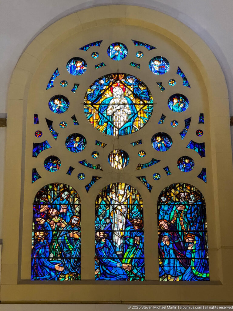 Arched stained-glass window with three figurative lower panels, a large central roundel, and many smaller blue and multicolored openings.