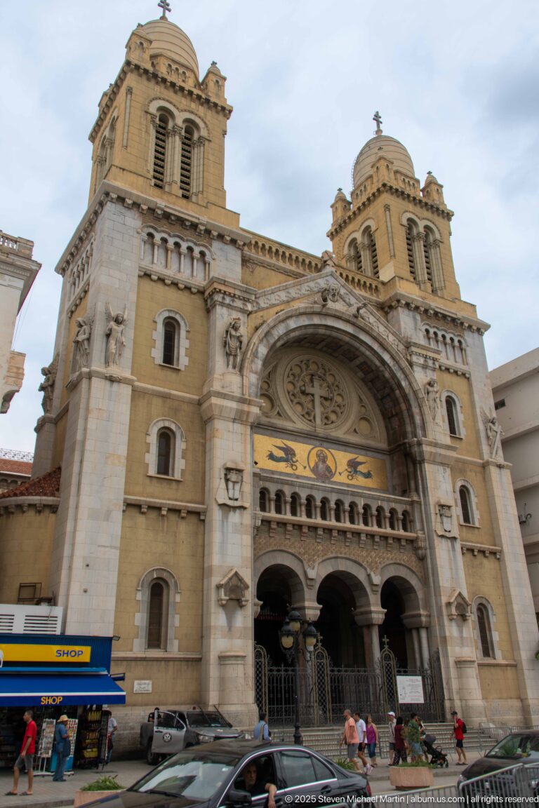 Main façade of St. Vincent de Paul Cathedral with twin bell towers, circular tracery, mosaic band, three arches, cars, and pedestrians.