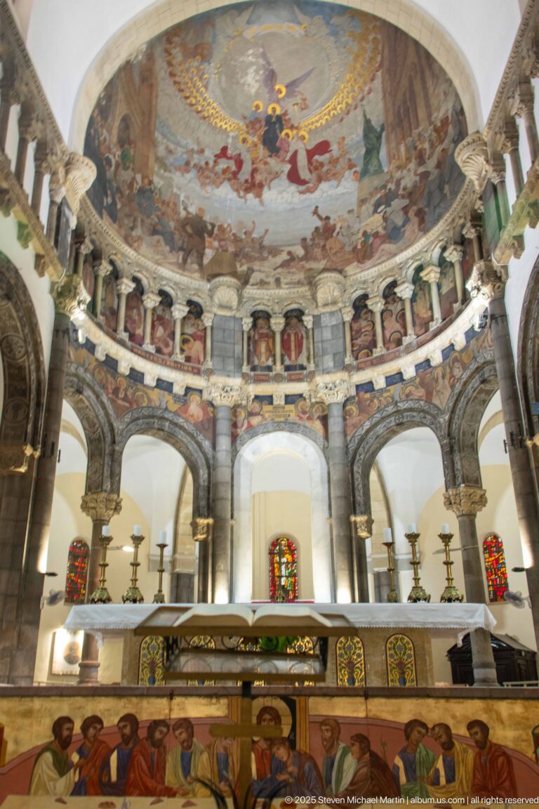 Cathedral sanctuary with altar, semicircular columned apse, stained glass, painted saints, and a large mural covering the apse vault.