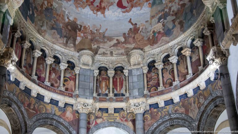 Curved cathedral apse with painted saints between ornate columns, inscriptions, decorative bands, and the lower portion of the vault mural.