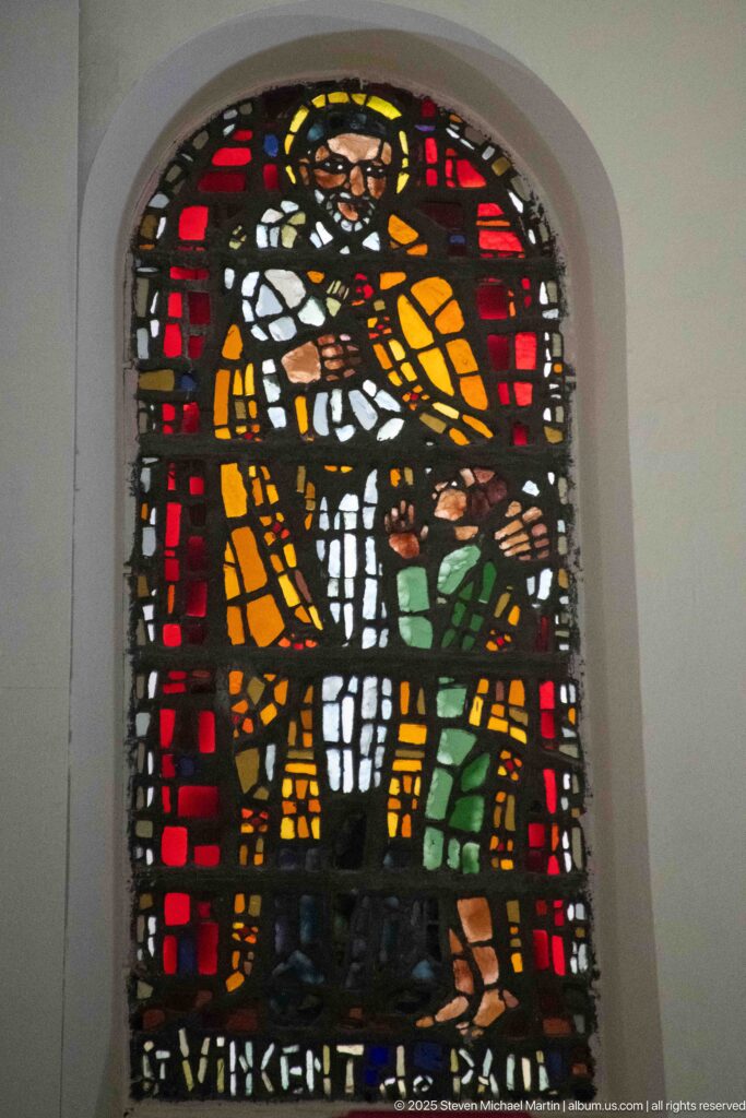 Small arched stained-glass panel with two haloed figures in orange and green against a red and dark background.