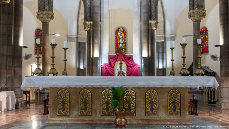 Frontal high altar with floral decorative panels, tall candlesticks, stained-glass windows, stone columns, magenta drapery, and a potted palm.