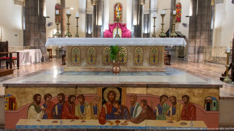 Cathedral altar above a painted Last Supper scene, with Christ and apostles below floral panels, candlesticks, stained glass, and drapery.