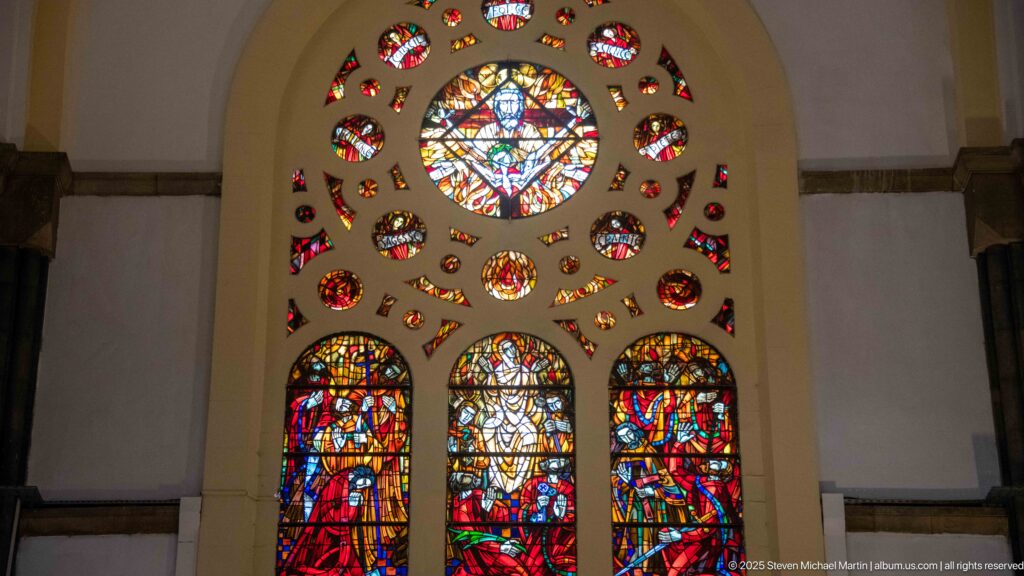 Warm red and amber stained-glass window with three figurative lower panels, central roundel, and many smaller colored openings.