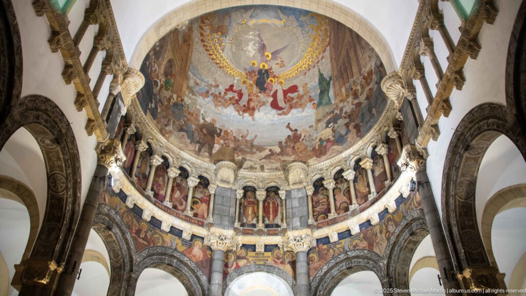 Wide interior view of the cathedral apse with monumental mural, painted saints, columns, and dark stone arches framing the sanctuary.