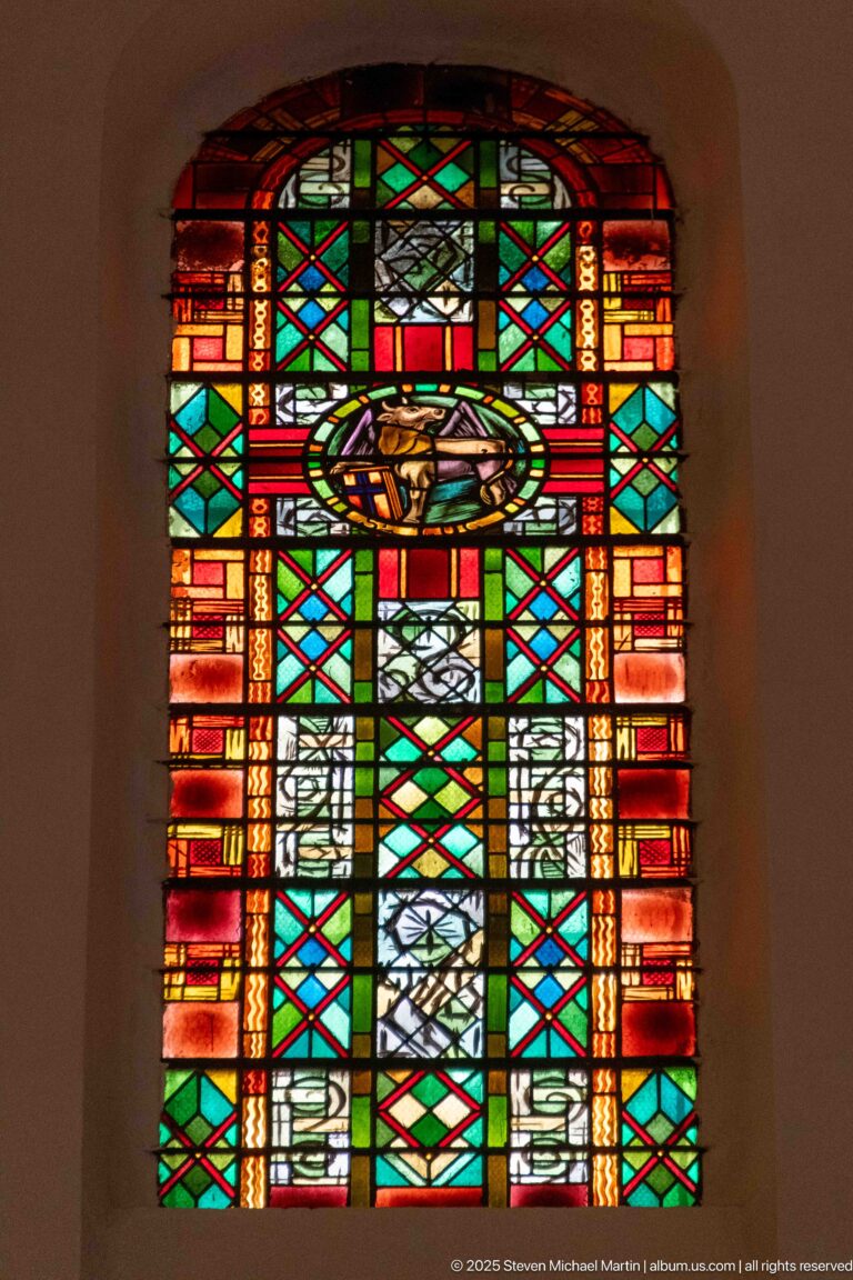 Narrow arched stained-glass window with irregular multicolored geometric panes, a vertical cross motif, and central oval element.